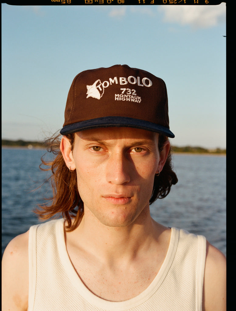 Man wearing Tombolo brown cap with a navy blue brim. The front features white embroidered text that reads “TOMBOLO” with a seashell illustration forming the letter “T,” and below it, “732 MONTAUK HIGHWAY.”