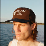 Man wearing Tombolo brown cap with a navy blue brim. The front features white embroidered text that reads “TOMBOLO” with a seashell illustration forming the letter “T,” and below it, “732 MONTAUK HIGHWAY.”