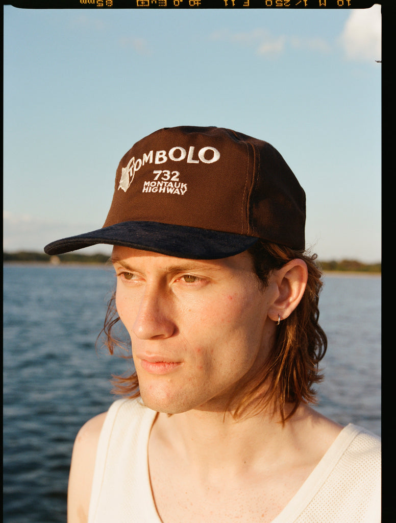 Man wearing Tombolo brown cap with a navy blue brim. The front features white embroidered text that reads “TOMBOLO” with a seashell illustration forming the letter “T,” and below it, “732 MONTAUK HIGHWAY.”