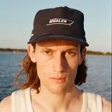 Male model with shoulder length brown hair wearing the 'Boston Whaler' 5-Panel Souvenir cap and a white tank top, shot on a background of water, a thin strip of land, and blue skies.