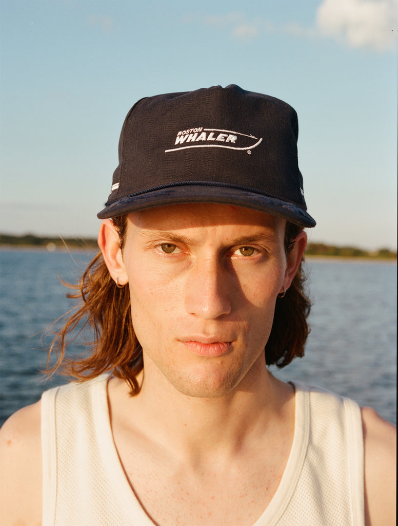 Male model with shoulder length brown hair wearing the 'Boston Whaler' 5-Panel Souvenir cap and a white tank top, shot on a background of water, a thin strip of land, and blue skies.