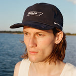 Male model with shoulder length brown hair wearing the 'Boston Whaler' 5-panel souvenir cap with a white tank top, shot on a background of the water, thin strip of land, and blue skies.