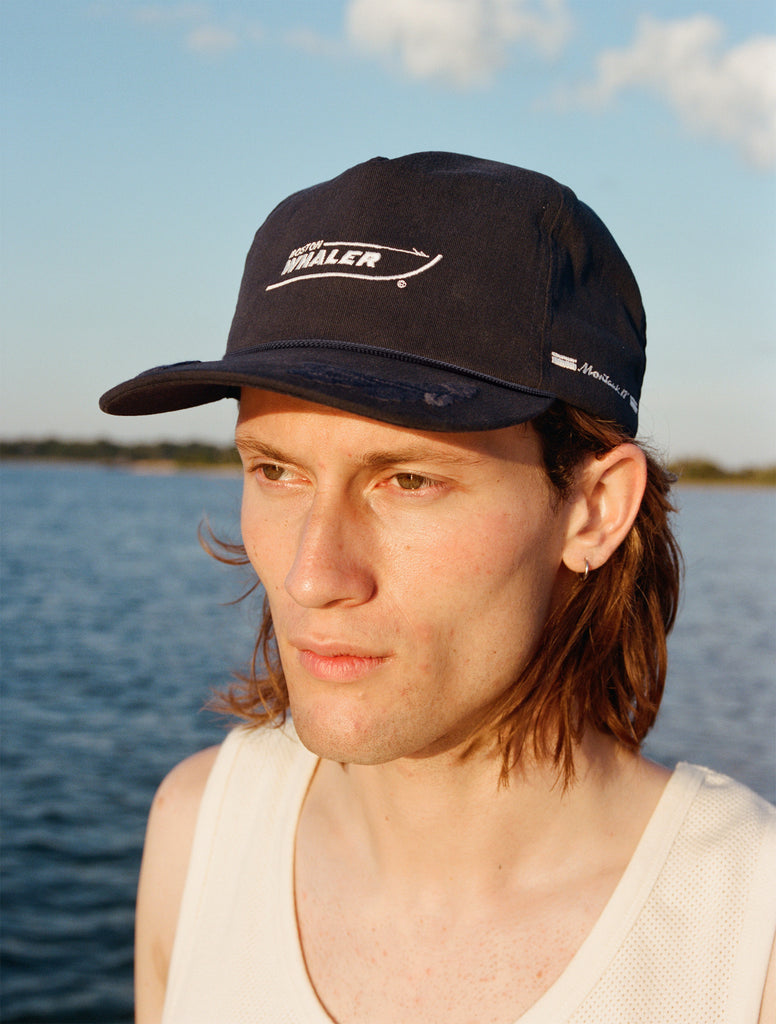Male model with shoulder length brown hair wearing the 'Boston Whaler' 5-panel souvenir cap with a white tank top, shot on a background of the water, thin strip of land, and blue skies.
