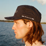 Male model with shoulder length brown hair wearing the 'Boston Whaler' 5-Panel Souvenir Cap while faced to the side, showcasing the 'Montauk '17' embroidery on the left panel, shot on a background of the water, a thin strip of land, and blue skies.