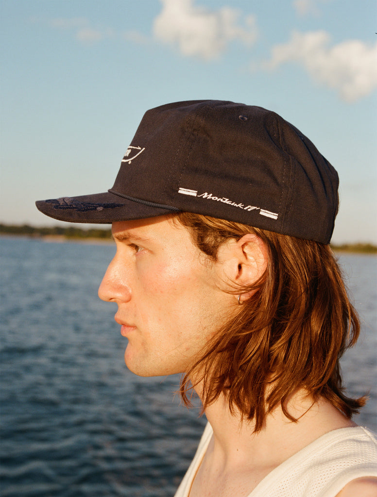 Male model with shoulder length brown hair wearing the 'Boston Whaler' 5-Panel Souvenir Cap while faced to the side, showcasing the 'Montauk '17' embroidery on the left panel, shot on a background of the water, a thin strip of land, and blue skies.