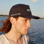 Male model with shoulder length brown hair wearing the 'Boston Whaler' 5-Panel Souvenir Cap wearing a white tank top faced to the side, shot on a background of the water, a thin strip of land, and blue skies.