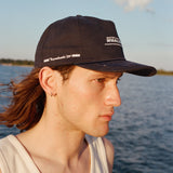 Male model with shoulder length brown hair wearing the 'Boston Whaler' 5-Panel Souvenir Cap wearing a white tank top faced to the side, shot on a background of the water, a thin strip of land, and blue skies.