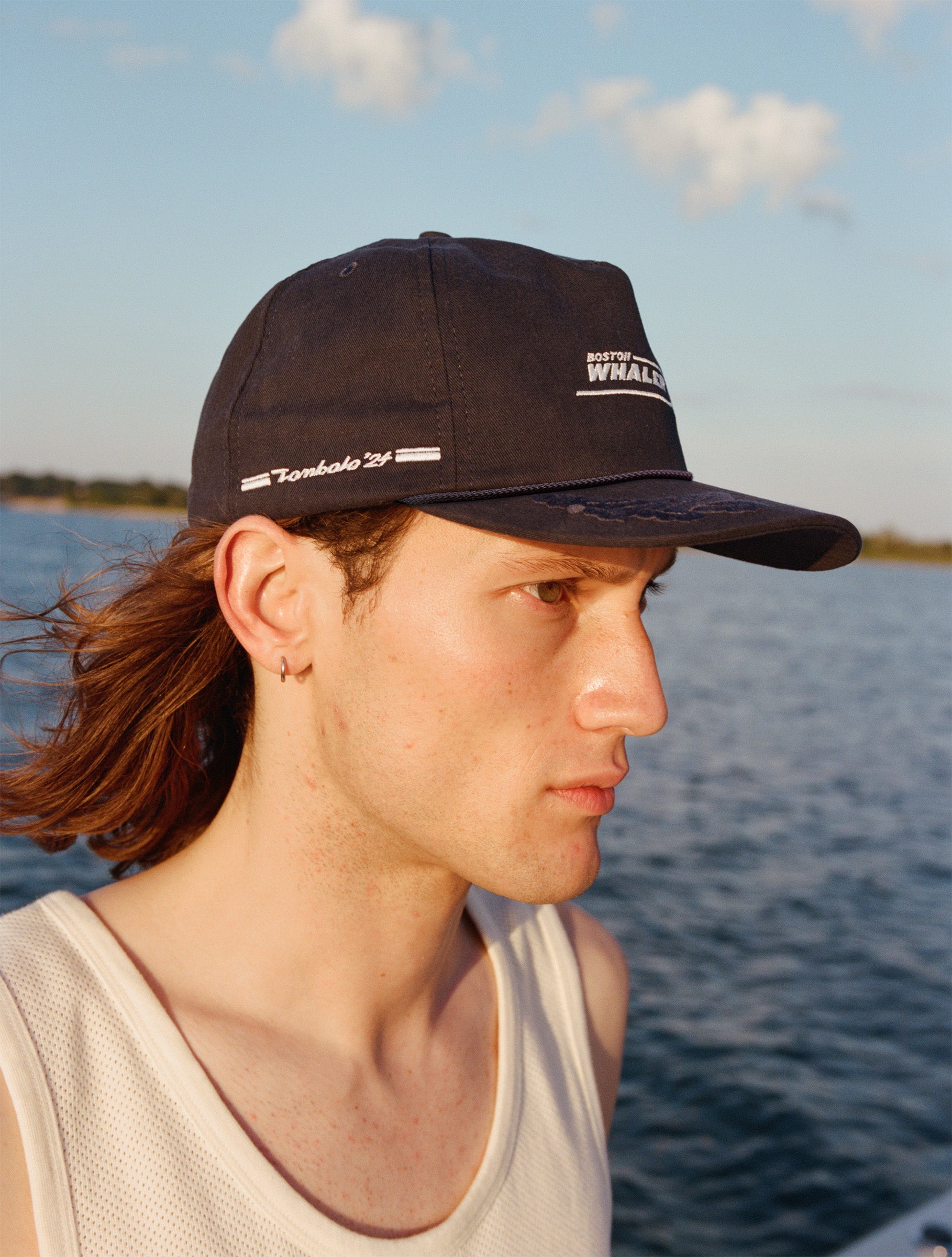 Male model with shoulder length brown hair wearing the 'Boston Whaler' 5-Panel Souvenir Cap wearing a white tank top faced to the side, shot on a background of the water, a thin strip of land, and blue skies.