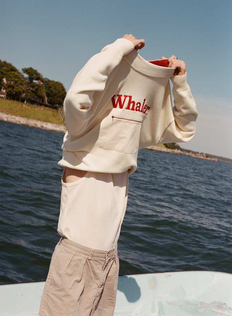 Male model in the process of taking off his 'Boston Whaler' Sweater, shot on a boat on the water with blue skies and bit of land visible.