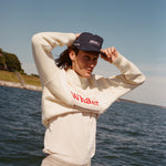 Male model with shoulder length brown hair wearing the 'Boston Whaler' 5-Panel Souvenir Cap with a white sweater and shirt, and khaki pants, using both hands to put on the cap, shot on a background of a boat on the water, a strip of land, and blue skies