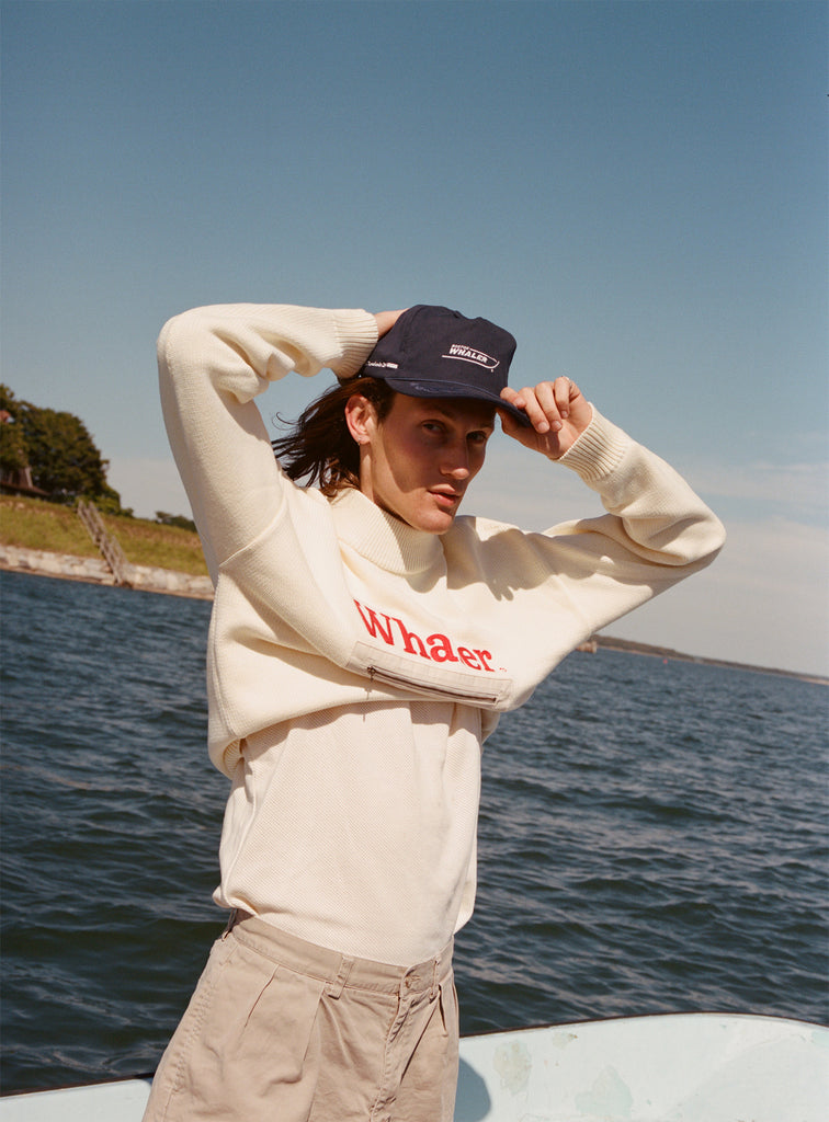 Male model with shoulder length brown hair wearing the 'Boston Whaler' 5-Panel Souvenir Cap with a white sweater and shirt, and khaki pants, using both hands to put on the cap, shot on a background of a boat on the water, a strip of land, and blue skies
