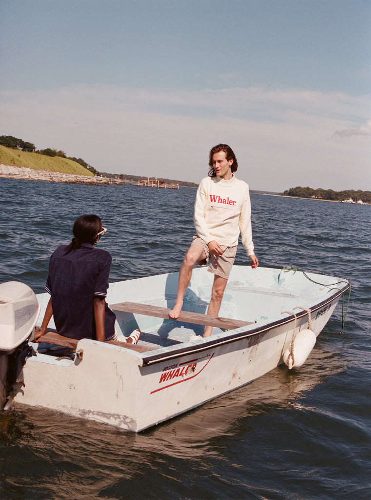 Male model standing on a boston whaler wearing his 'boston whaler' sweater and khaki shorts while talking to a seated female model wearing the 'boston whaler' cabana in navy.
