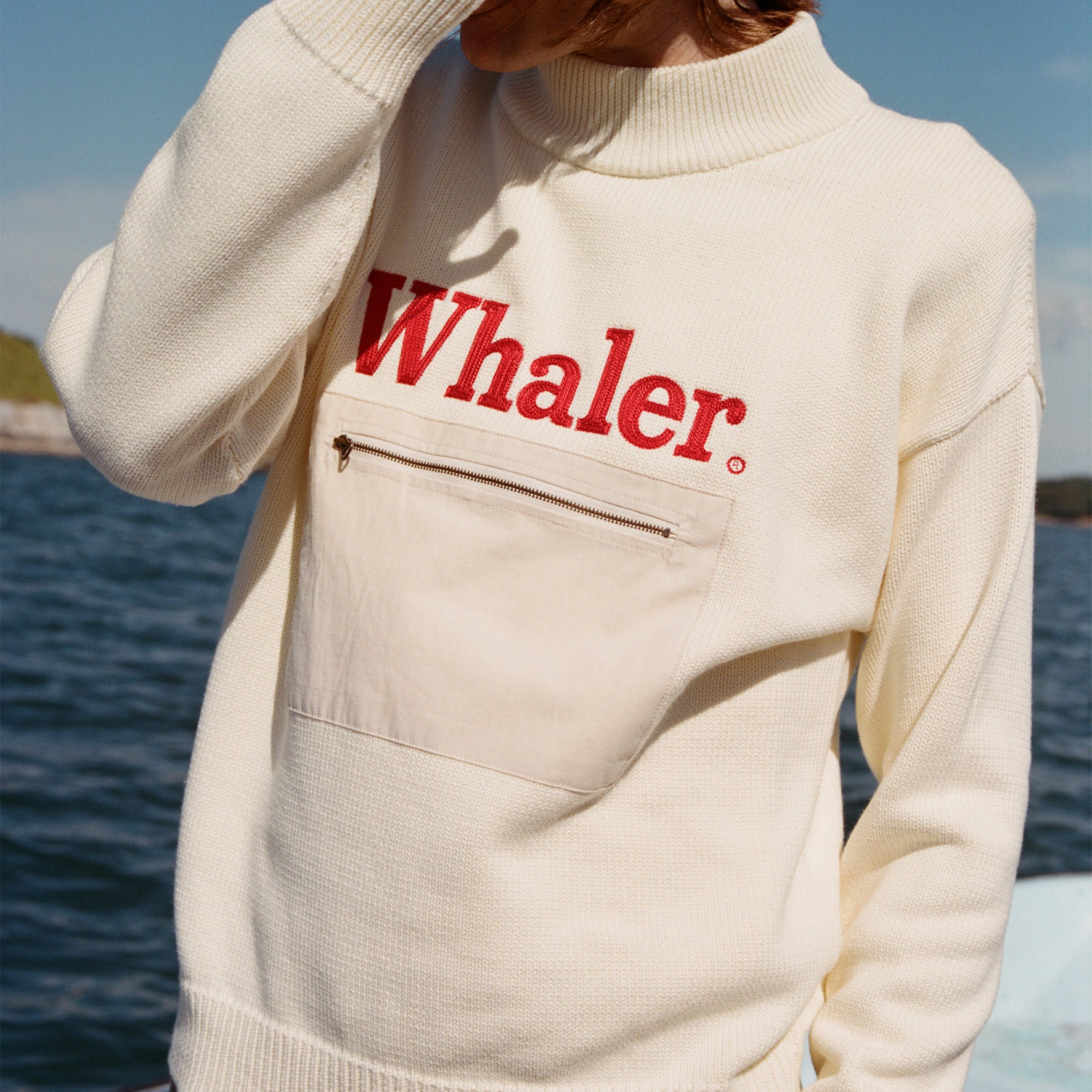 Male model with shoulder length brown hair wearing the 'Boston Whaler' sweater, shot on a background of a boat, water, and blue skies.