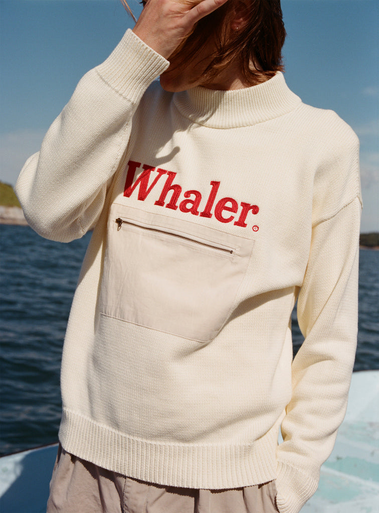 Male model with shoulder length brown hair wearing the 'Boston Whaler' sweater, shot on a background of a boat, water, and blue skies.