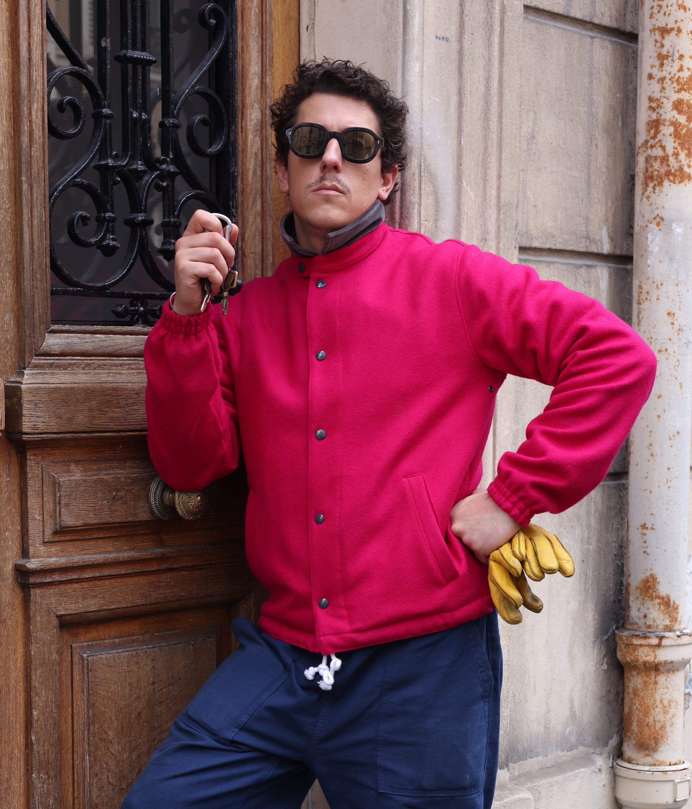 Young Male model wearing Lesca Lunetier X Tombolo Lenses alongside a bright red button up wool jacket while holding keys and leaning against a wooden door.