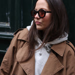Young female model wearing Lesca Lunetier X Tombolo sunglasses with black frames and coral colored lenses in addition to a brown unbottoned trench coat with a grey hoodie while leaning against a forest green background.