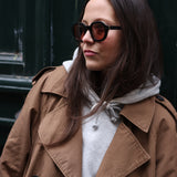 Young female model wearing Lesca Lunetier X Tombolo sunglasses with black frames and coral colored lenses in addition to a brown unbottoned trench coat with a grey hoodie while leaning against a forest green background.