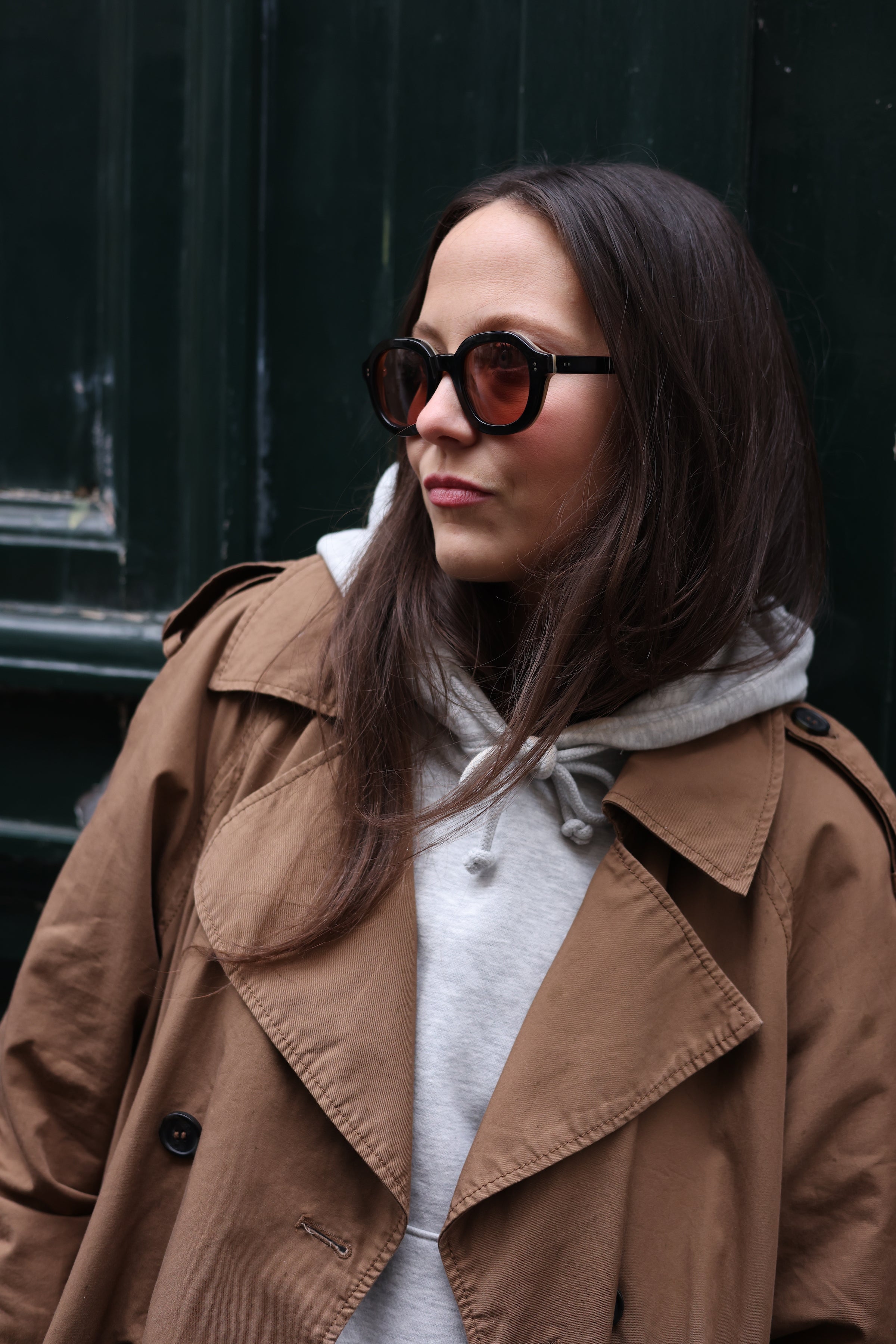 Young female model wearing Lesca Lunetier X Tombolo sunglasses with black frames and coral colored lenses in addition to a brown unbottoned trench coat with a grey hoodie while leaning against a forest green background.
