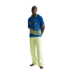 Man wearing a blue and yellow beach towel top with light green pants.