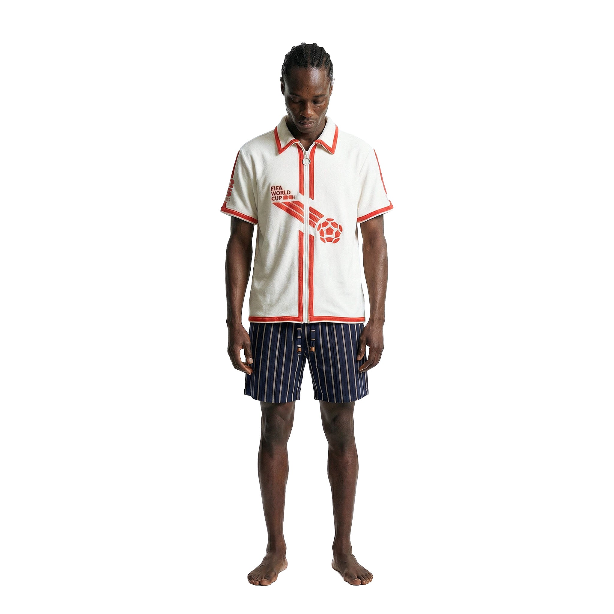 Model wearing a white terry cloth short-sleeve shirt with red trim and red FIFA World Cup 26™ graphics, paired with navy striped shorts, shown against a white background.