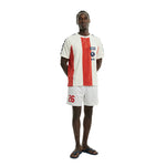 Male model wearing a white terry cloth short-sleeve shirt with wide vertical red stripes, a quarter-zip neckline, and FIFA World Cup patches, paired with white shorts, shown against a white background.