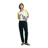 Female model wearing a white terry cloth shirt with yellow raglan sleeves, a black-and-white striped ribbed collar, a quarter-zip neckline, and a black Statue of Liberty graphic on the front, paired with black pants.