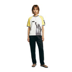 Female model wearing a white terry cloth shirt with yellow raglan sleeves, a black-and-white striped ribbed collar, a quarter-zip neckline, and a black Statue of Liberty graphic on the front, paired with black pants.