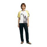 Female model wearing a white terry cloth shirt with yellow raglan sleeves, a black-and-white striped ribbed collar, a quarter-zip neckline, and a black Statue of Liberty graphic on the front, paired with black pants.