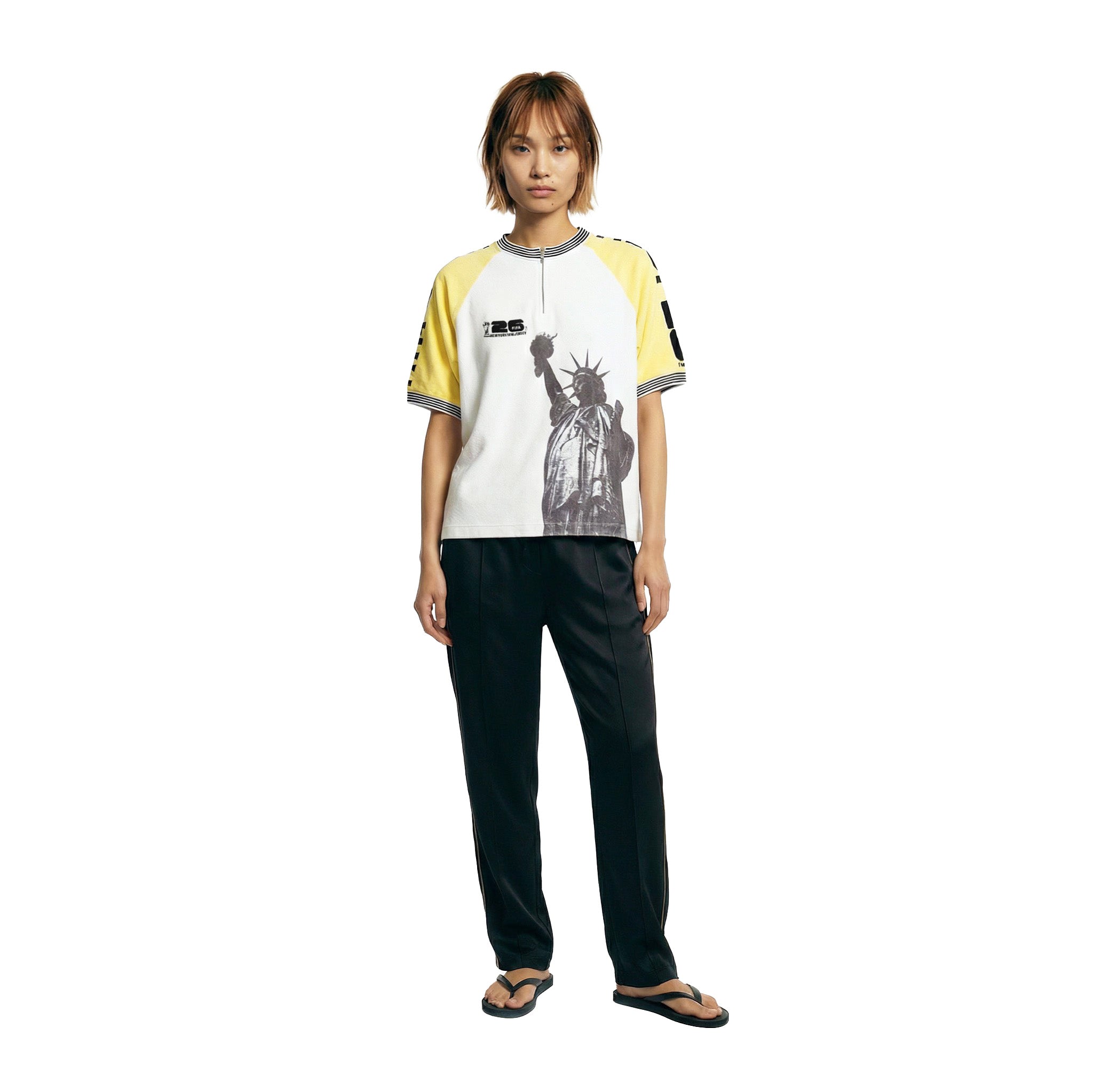 Female model wearing a white terry cloth shirt with yellow raglan sleeves, a black-and-white striped ribbed collar, a quarter-zip neckline, and a black Statue of Liberty graphic on the front, paired with black pants.