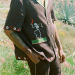 Close up view of body of man standing in field wearing full 'Picante' cabana set in linen. Embroidered skeleton on shirt is clearly visible.
