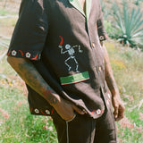 Close up view of body of man standing in field wearing full 'Picante' cabana set in linen. Embroidered skeleton on shirt is clearly visible.