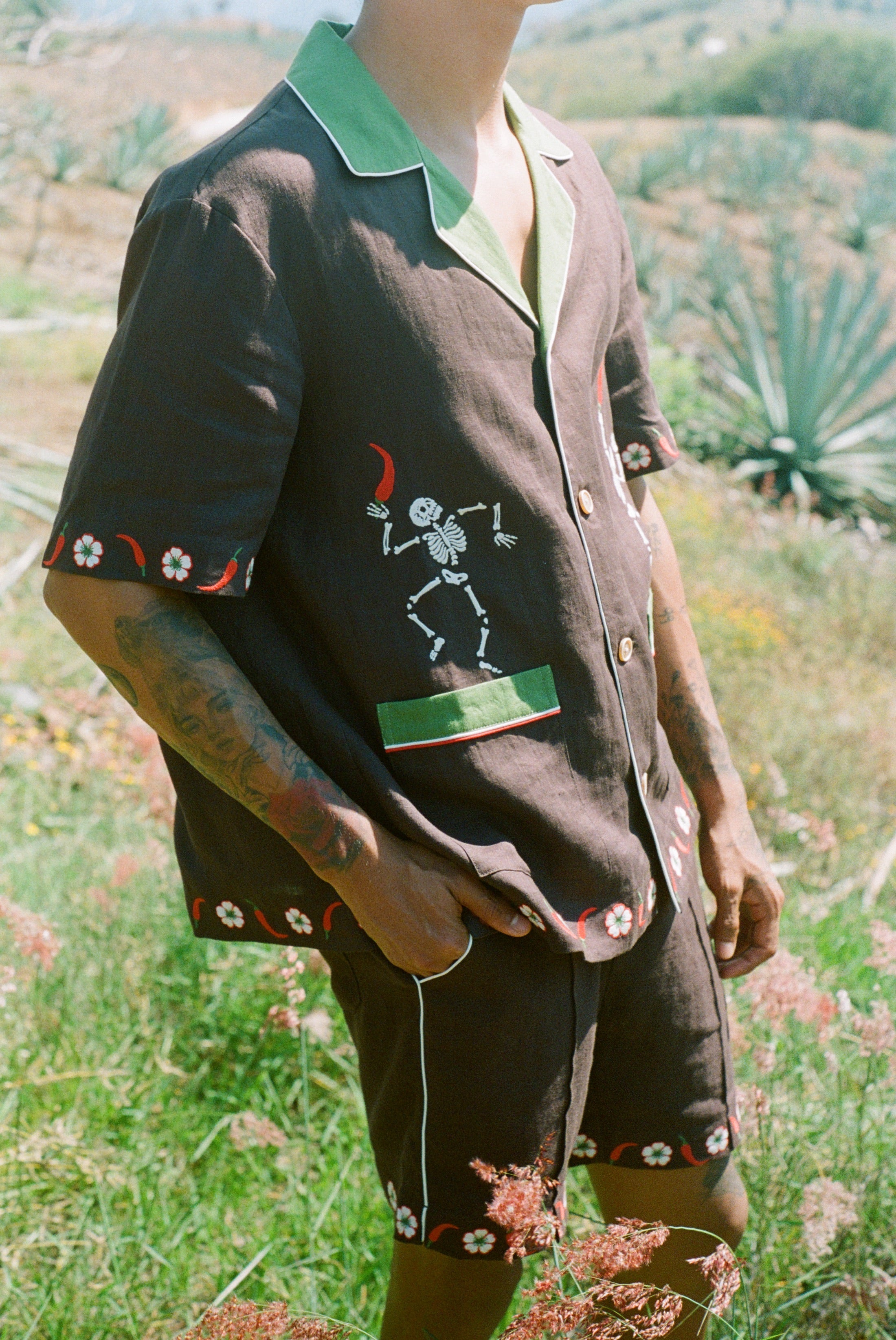 Close up view of body of man standing in field wearing full 'Picante' cabana set in linen. Embroidered skeleton on shirt is clearly visible.