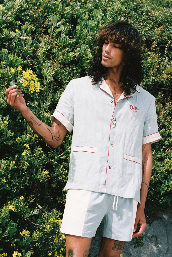 man holds leaves from background wearing linen shirt and matching oyster trunks