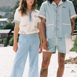 woman stares at camera wearing Tombolo shirt and man stares at camera wearing blue Tombolo shirt and swim trunks