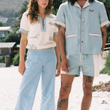 woman stares at camera wearing Tombolo shirt and man stares at camera wearing blue Tombolo shirt and swim trunks
