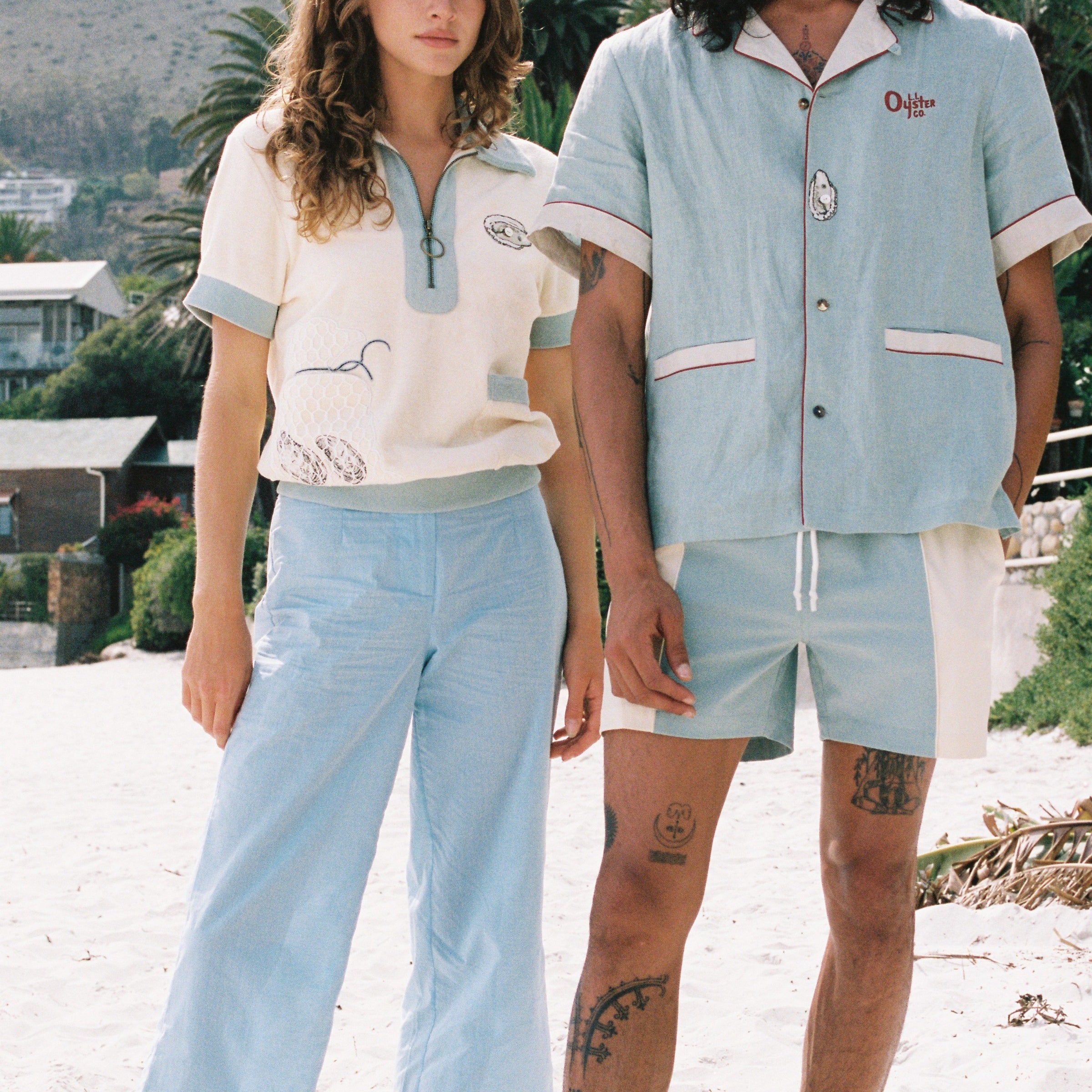 woman stares at camera wearing Tombolo shirt and man stares at camera wearing blue Tombolo shirt and swim trunks
