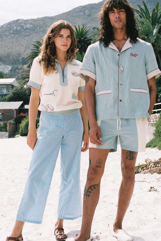 woman stares at camera wearing Tombolo shirt and man stares at camera wearing blue Tombolo shirt and swim trunks