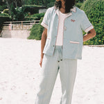 male model wearing blue linen tombolo pants and Tombolo Billion Oyster Project blue linen shirt while looking off to the left on a beach