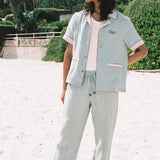 male model wearing blue linen tombolo pants and Tombolo Billion Oyster Project blue linen shirt while looking off to the left on a beach