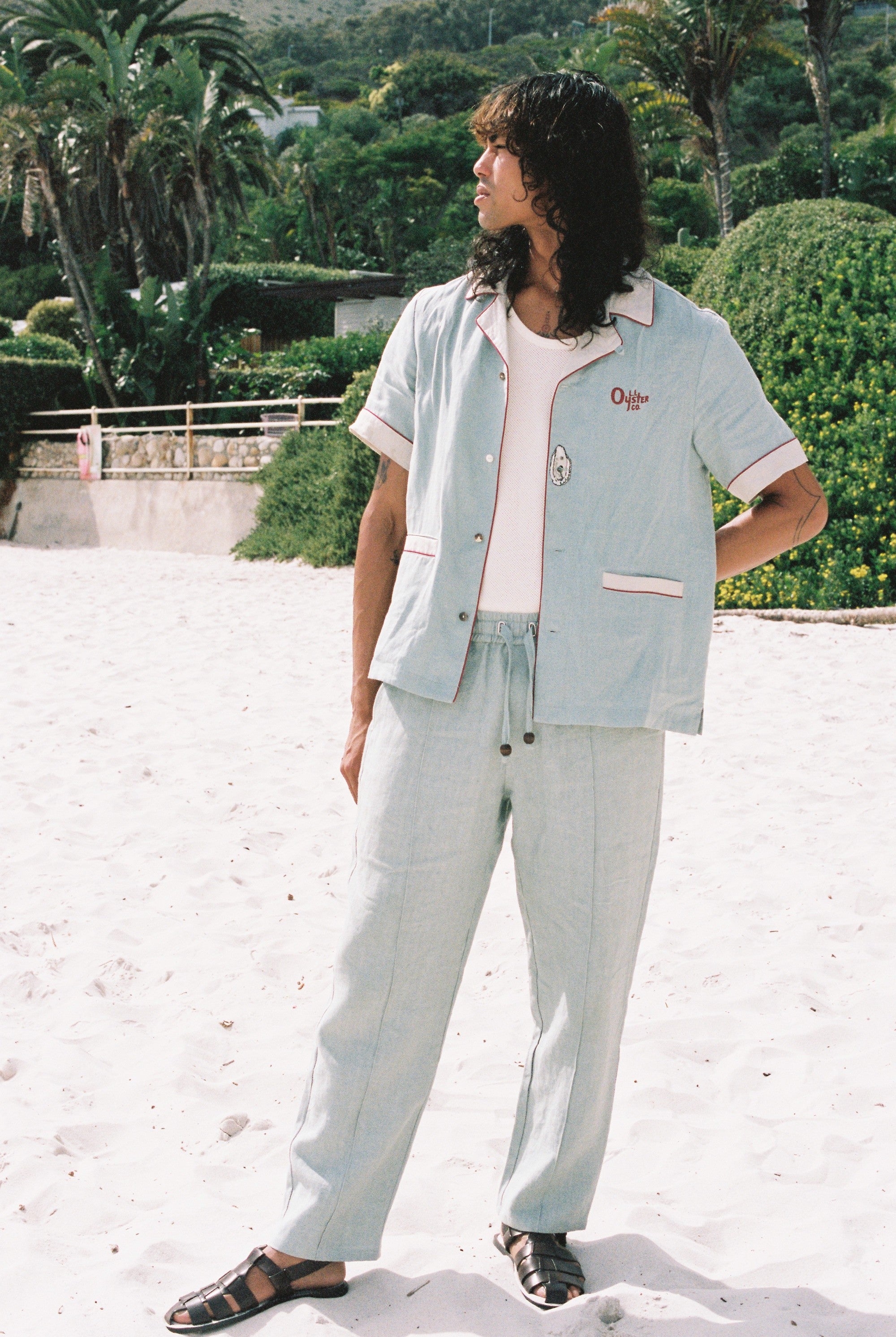 male model wearing blue linen tombolo pants and Tombolo Billion Oyster Project blue linen shirt while looking off to the left on a beach