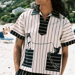 Man on the beach looking to the left, wearing beige Tombolo striped pop-over shirt and matching navy striped pants