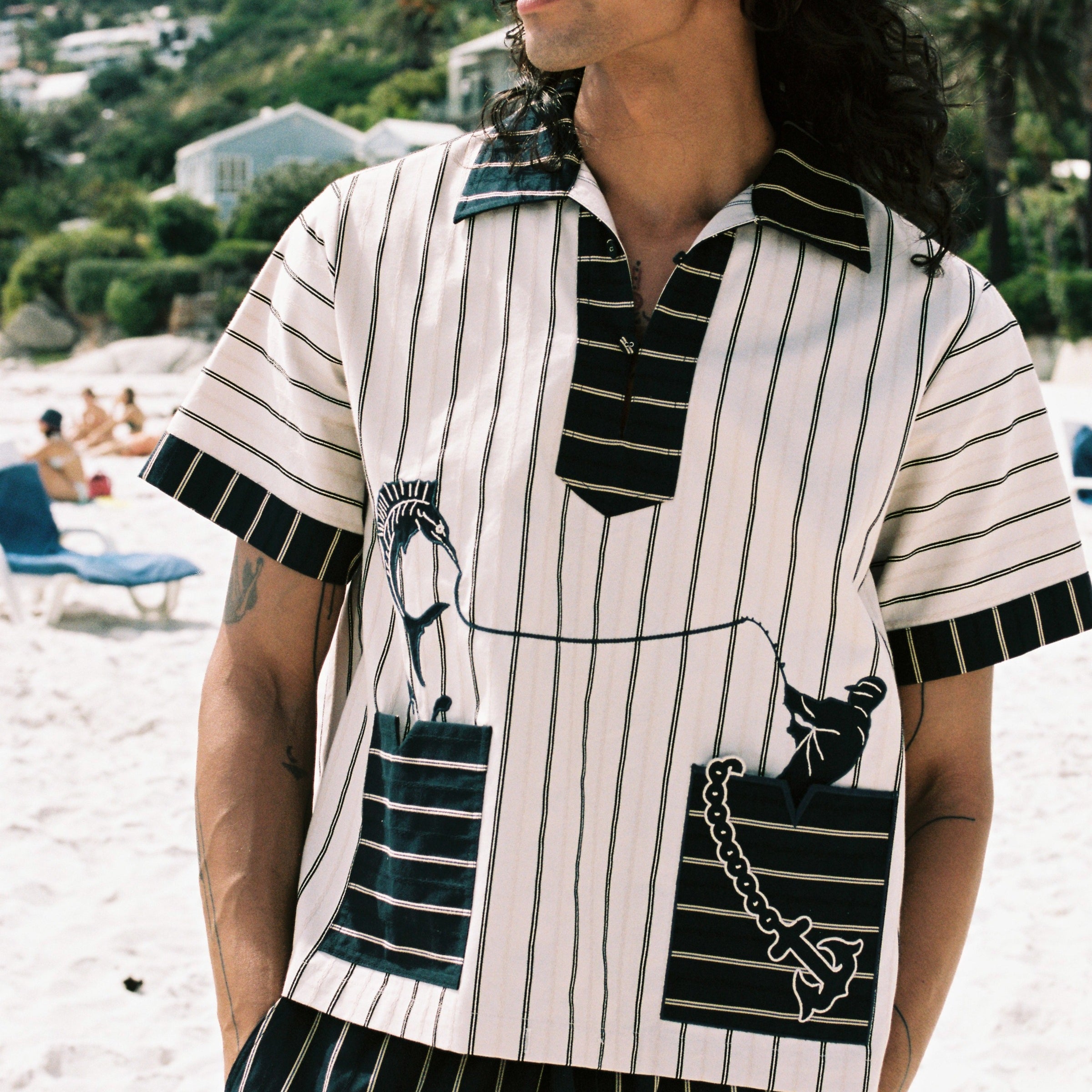 Man on the beach looking to the left, wearing beige Tombolo striped pop-over shirt and matching navy striped pants