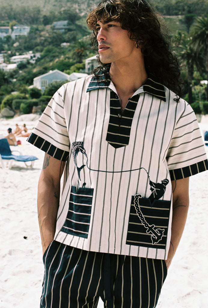 Man on the beach looking to the left, wearing beige Tombolo striped pop-over shirt and matching navy striped pants
