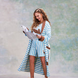Female model with long blonde hair looking down at a newspaper in her hands while wearing 'The Sopranos' Fowl Play Robe in light blue and white horizontal striping alongside matching Fowl Play shorts and shirt with Ugg Slippers.