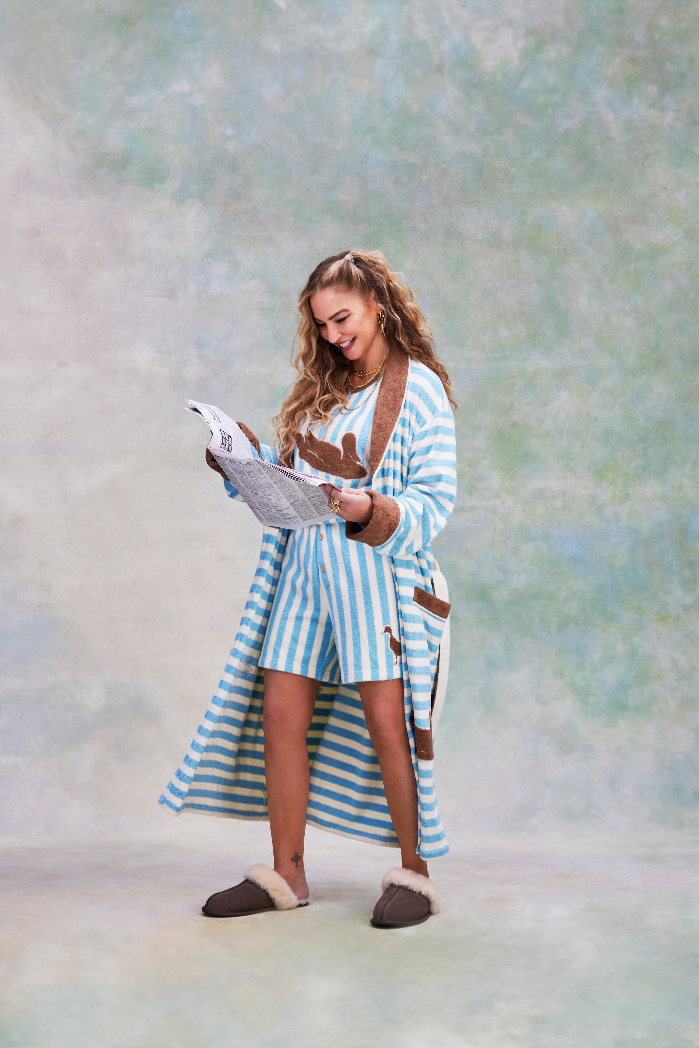 Female model with long blonde hair holding a newspaper while wearing 'The Sopranos' Fowl Play shorts in light blue and white stripes with matching Fowl Play Top and Robe in addittion to Ugg Slippers, shot a textured white background.