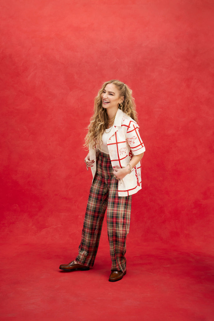 Female model with long blonde hair wearing 'The Sopranos' Satriale's (Gabagool) unbuttoned with plaid pants, brown loafers, laughing, against a red background.