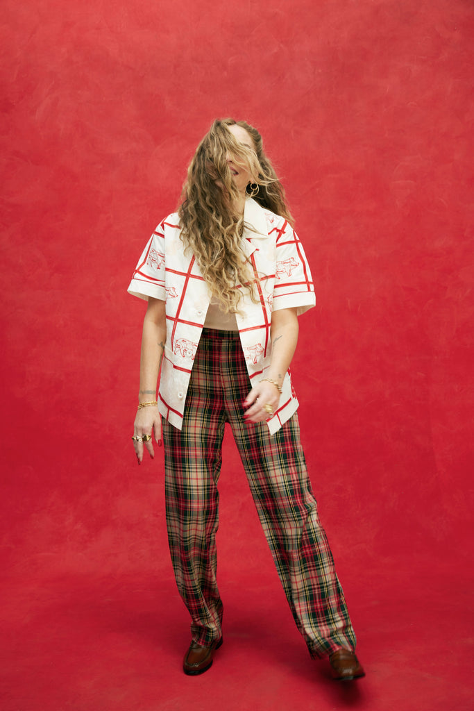 Female model with long blonde hair in plaid pants and brown loafers wearing an unbuttoned 'The Sopranos' Satriale's (Gabagool) with a white shirt underneath, shot on a red background.