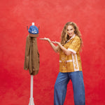 Female model with long blond hair wearing 'The Sopranos' Satriale's (Pruh-Zhoot) while using her hands to showcase a Houndstooth blazer hanging, alongside a hat hanging, while the model wears baggy jeans and brown loafers, all shot on a red background.
