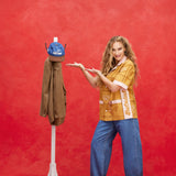 Female model with long blond hair wearing 'The Sopranos' Satriale's (Pruh-Zhoot) while using her hands to showcase a Houndstooth blazer hanging, alongside a hat hanging, while the model wears baggy jeans and brown loafers, all shot on a red background.
