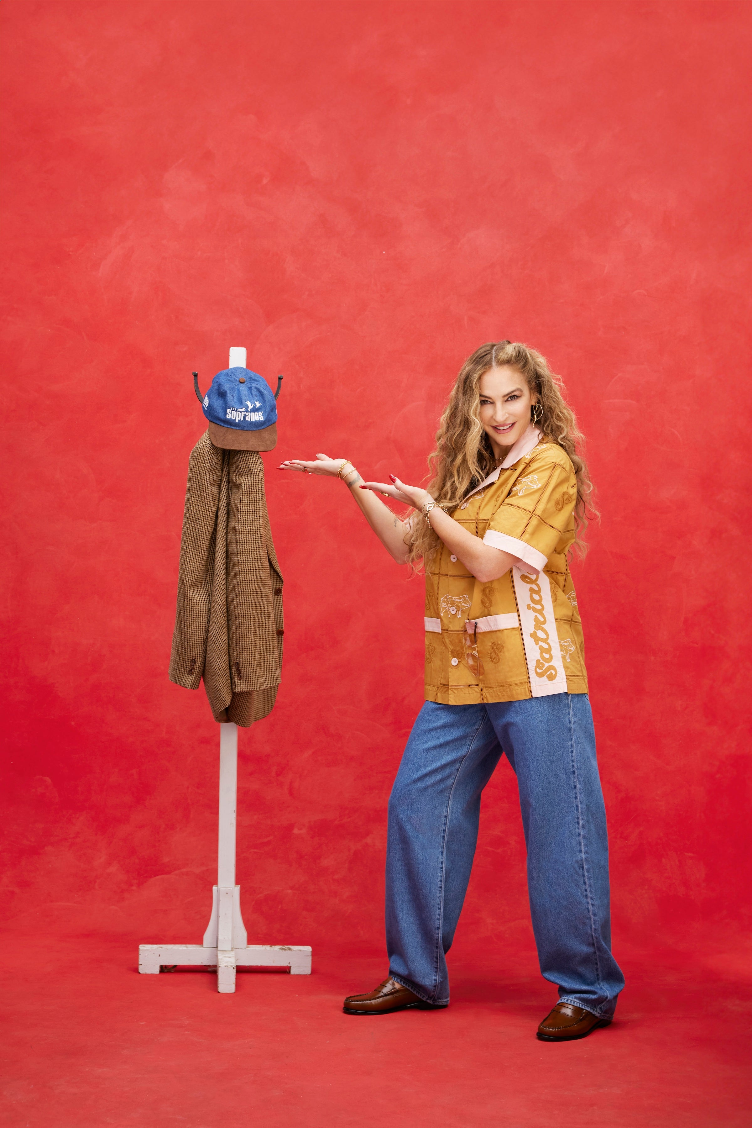 Female model with long blonde hair standing beside a coat rack holding a Houndstooth blazer and 'The Sopranos' Fowl Play 6-Panel Souvenir Cap, while the model wears a Satriale's 'Pruh-Zhoot' Caban with long flowey jeans and brown loafers, shot on a red textured background.